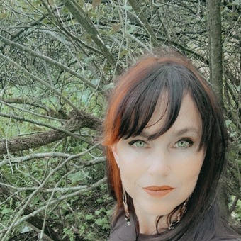 Photo of Amy McCoy with a shrub or tree, long earrings, black hair with wispy layered bangs and a red streak, and a subtle smile.