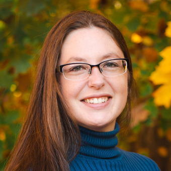 Photo of Briana Kangas Otten, wearing a dark blue turtle neck sweater rectangle-framed glasses, long red-brown hair styled down, smiling.