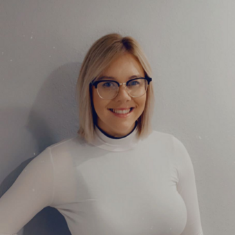 Photo of Brittany Bartley, white turtleneck, shoulder length blonde hair styled down, square-framed glasses, smiling.