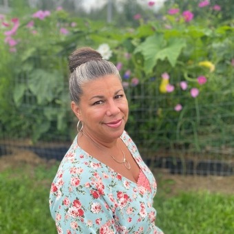 Phot of Carrie Perez, in front of tall flowers, light blue blouse or dress with pink floral patterns, silver earrings and necklace, dark gray hair styled up in a bun, smiling.