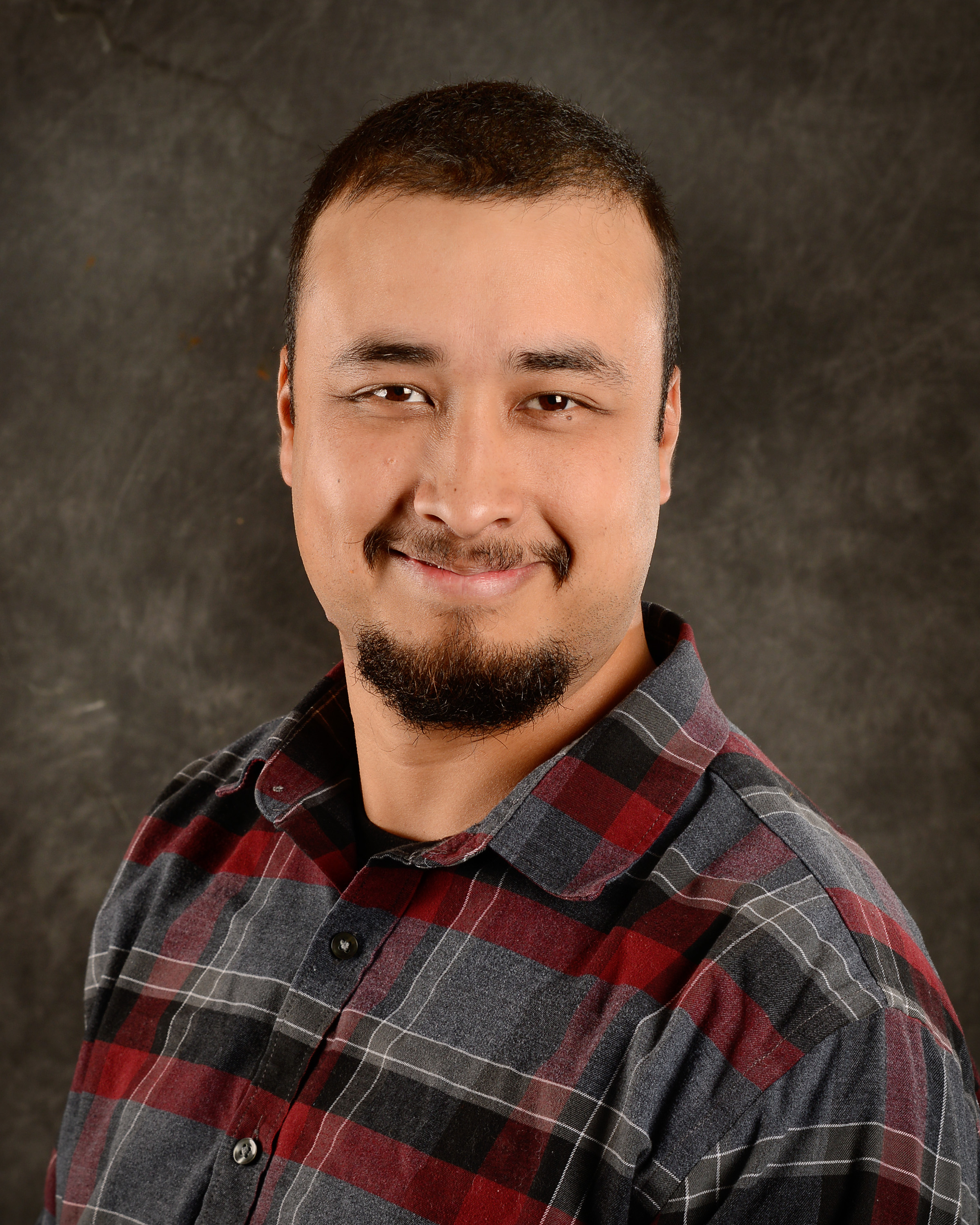 Photo of Jason LeBlanc Jr., wearing a black-grey-red plaid shirt, short dark brown hair, smiling with a royale beard.