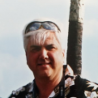 Photo of John Adair, outside, sunglasses resting atop head over gray hair, black with white floral print dress t-shirt, side-lit, smiling.