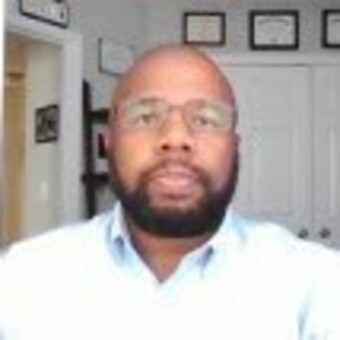 Photo of Ty Adams, framed degrees on walls in the background, light blue dress shirt, rectangle-framed glasses, hairstyle is shaved with short boxed beard, with a subtle smile.