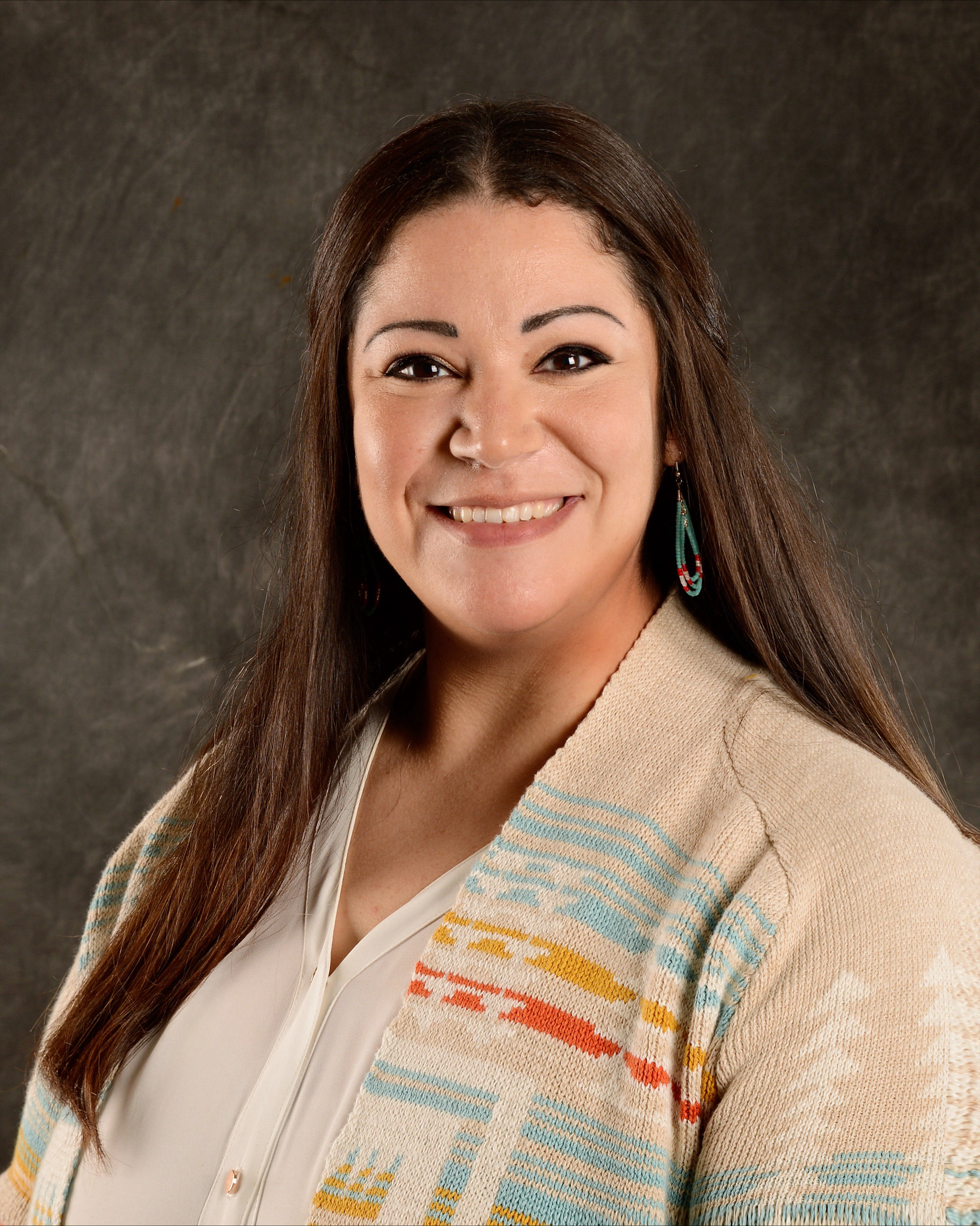 Photo of Candice LeBlanc, white blouse with light Pendleton pattern cardigan, long beaded earrings, long brown hair styled partially up, smiling.