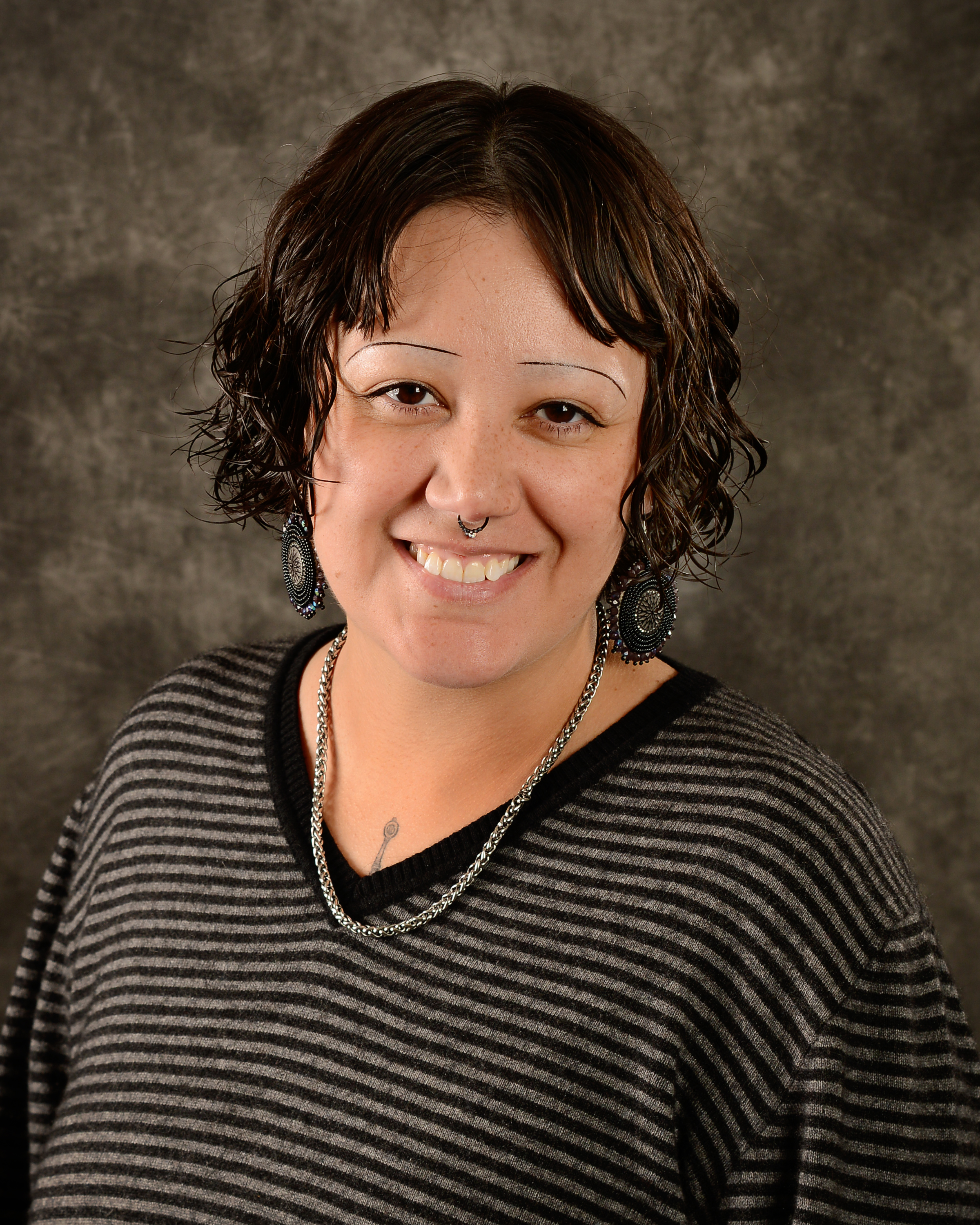 Photo of Chelsey Cameron, painting of an eagle feather on the wall behind, dark brown hair in a pony tail over the left shoulder, round beaded earrings, black dress shirt, septum hoop, smiling.