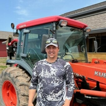 Dave Corey wearing a patterned long-sleeve shirt, a hat with sunglasses tucked on the brim, standing in frone of an orange tractor with green grass in the background, next to the Waishkey Bay Farm offices.
