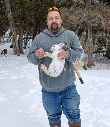 Dennis Carrick in jeans and a hoodie holding a rabbit and a snare. There is a pair of wire cutters tucked under arm, sunglasses on top of his head, and snow on the ground with trees behind him.